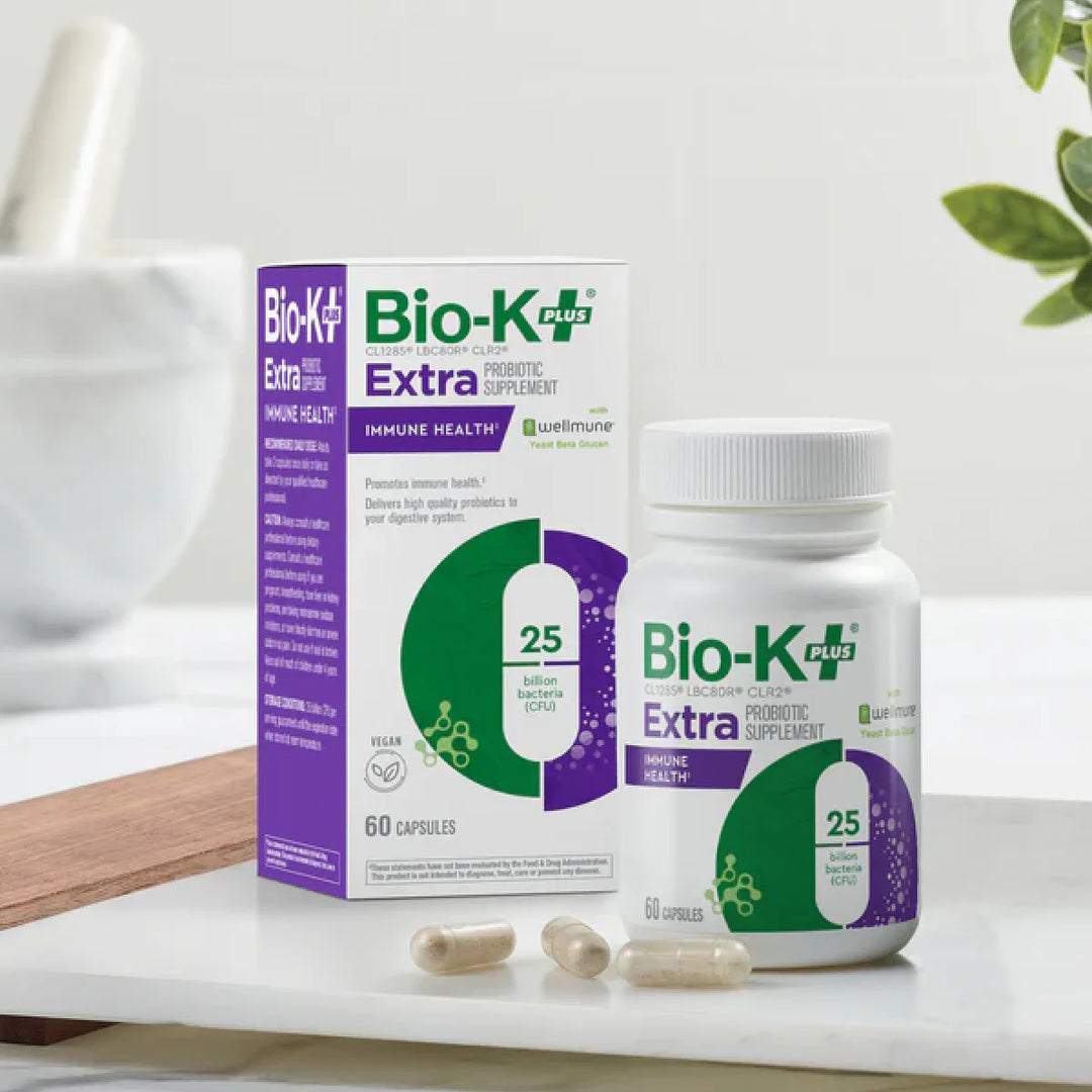 Bio-K+ Extra probiotic supplement bottle and packaging on a white surface with a plant in the background.