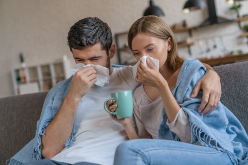 Man and woman sitting on a couch, both holding tissues and a mug, wrapped in a blanket.